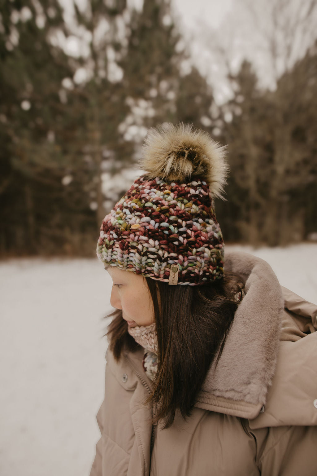 Person wearing a colorful knit beanie and beige coat in a snowy forest setting