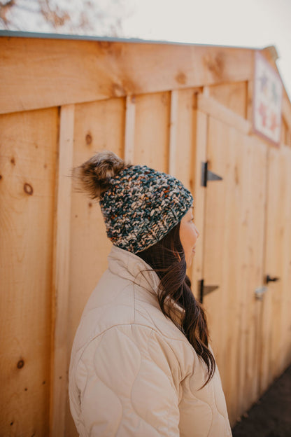 A woman with long dark hair and beige quilted jacket wears a colorful green beanie with faux fur pom pom. 