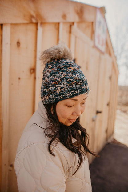 A person wearing a green merino wool knit hat with a faux fur pom pom and beige quilted jacket.