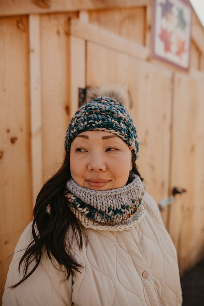 Woman with beige jacket wears a green colorful knit beanie and cowl. 