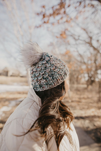 A woman wearing a blue and gray speckled merino wool knit hat with a faux fur pom pom.