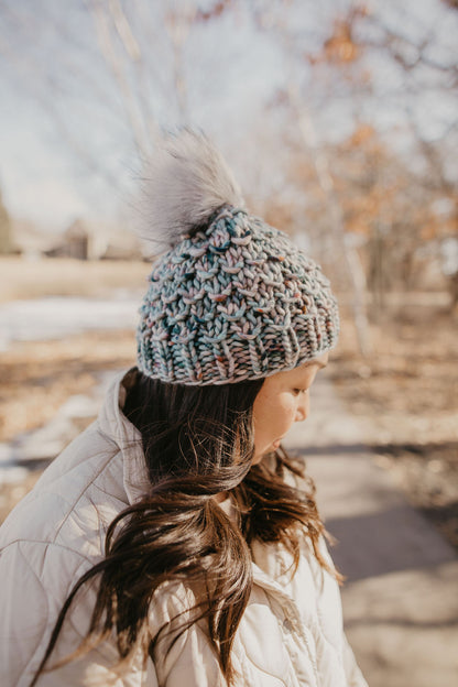 A woman wearing a blue and gray speckled merino wool knit hat with a faux fur pom pom.