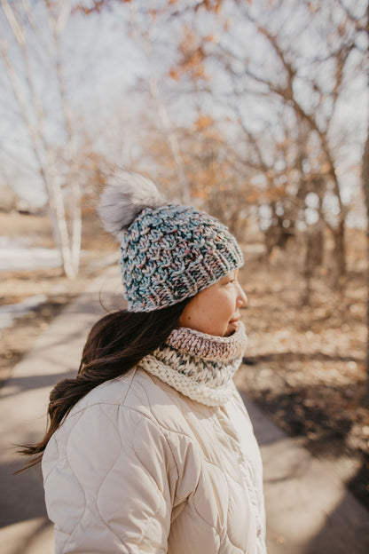 A woman wearing a beige jacket and a blue and gray speckled merino wool knit hat with a faux fur pom pom.