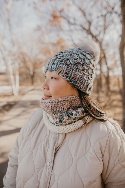 A woman wearing a blue and gray speckled merino wool knit hat with a faux fur pom pom