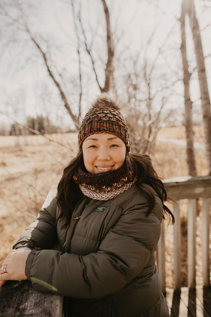 Woman wearing a dark green jacket wears a colorful brown knitted hat and cowl in a forest setting