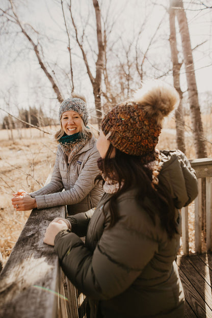 Two women wearing colorful knit hats talking to each other on a wooden deck in a forest setting
