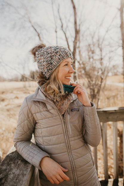 Woman wearing a blue merino wool hat and cowl with a gray jacket on a wooden dock in the forest