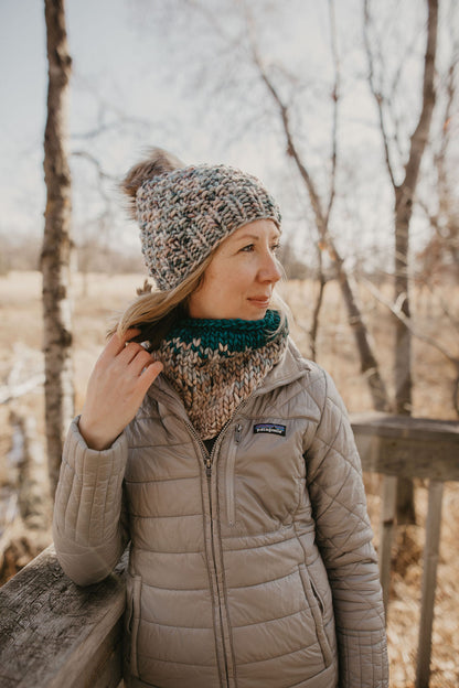 Woman wearing a blue merino wool hat and cowl with a gray jacket on a wooden dock in the forest