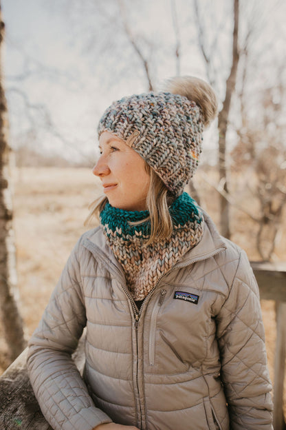 Woman wearing a blue merino wool hat and cowl with a gray jacket on a wooden dock in the forest