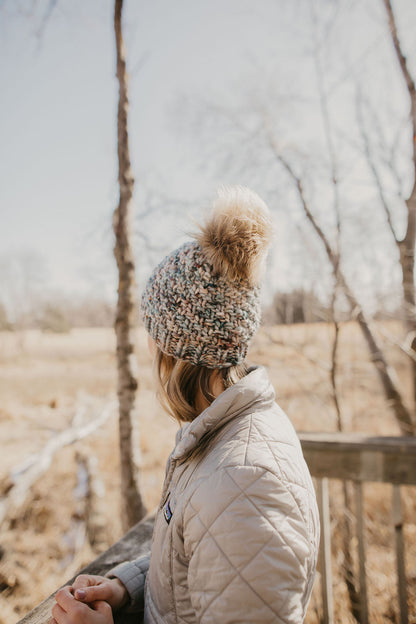 Woman wearing a blue merino wool hat and cowl with a gray jacket on a wooden dock in the forest