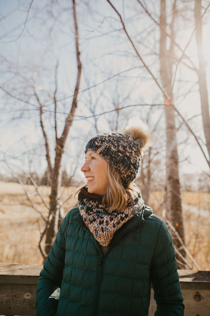 Woman wearing green multicolor knit hat with faux fur pom pom and knit cowl on a wooden deck in the forest