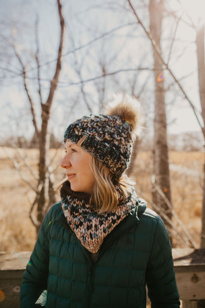 Woman wearing green multicolor knit hat with faux fur pom pom and knit cowl on a wooden deck in the forest