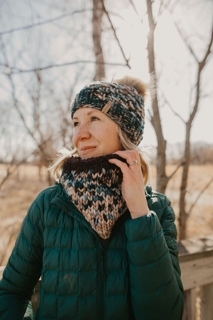 Woman wearing green multicolor knit hat with faux fur pom pom and knit cowl on a wooden deck in the forest