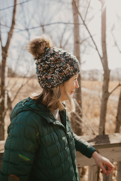 Woman wearing green multicolor knit hat with faux fur pom pom on a wooden deck in the forest