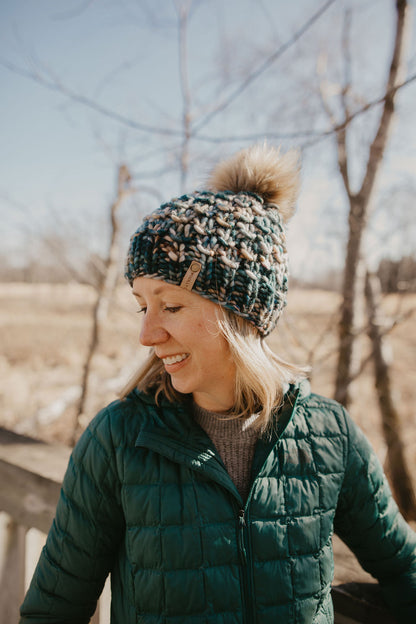 Woman wearing green multicolor knit hat with faux fur pom pom and knit cowl on a wooden deck in the forest