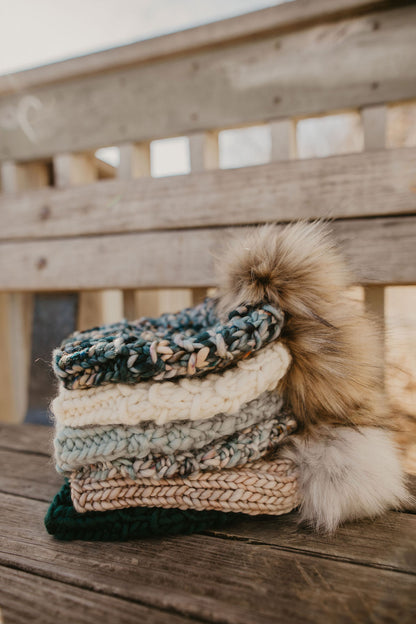 A stack of six colorful chunky knit hats with faux fur pom poms on a wooden deck