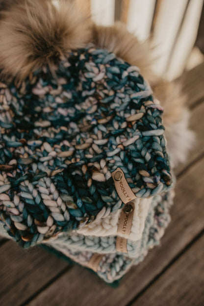 A stack of knit hats with fluffy pom poms sit on a wooden deck.
