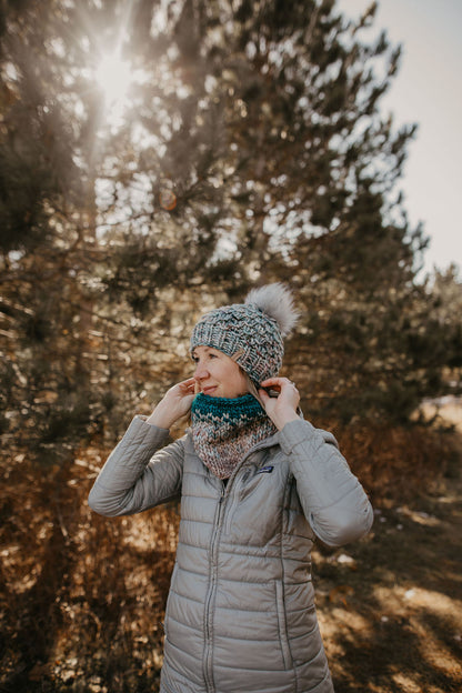 Woman wearing blue knitted hat and cowl with a quilted gray jacket in a forest setting