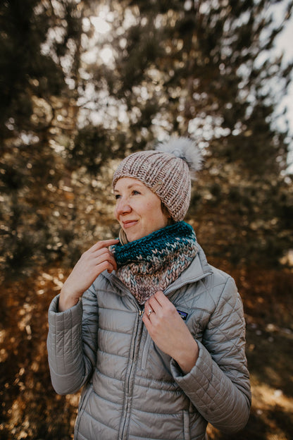 Woman in a gray puffer jacket wears a gray ribbed knit hat and scarf in a forest