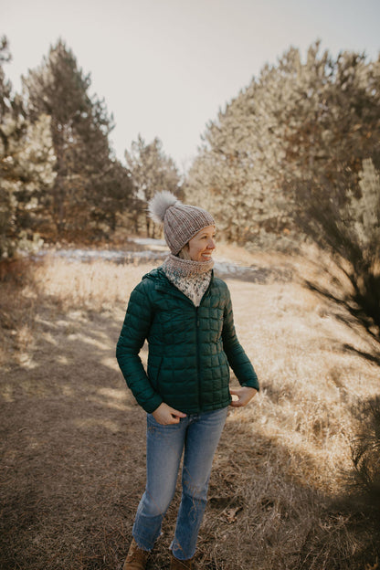Woman wearing a green puffer coat with a gray knit hat and scarf in a forest 