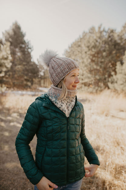 Woman wearing a green puffer coat with a gray knit hat and scarf in a forest 