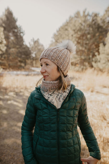 Woman wearing a green puffer coat with a gray knit hat and scarf in a forest 