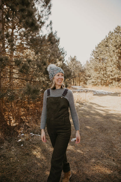 Woman wearing blue knitted beanie with faux fur pom pom and green wool overalls in a forest