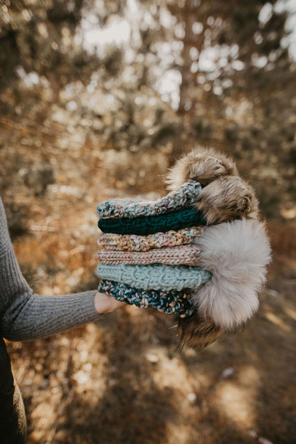 A person holds a stack of six hand knitted beanies with faux fur pom poms in a forest setting
