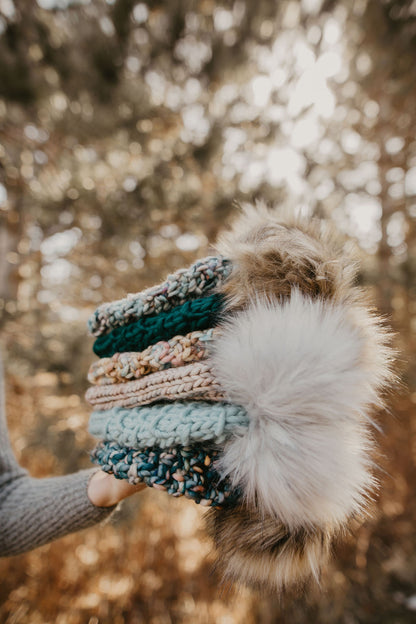 A person holds a stack of six chunky knit hats with fluffy faux fur pom poms in a forest setting