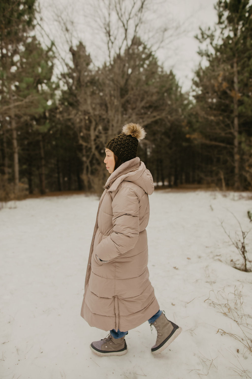 Person walking in a snowy forest wearing a long beige coat and knitted hat.
