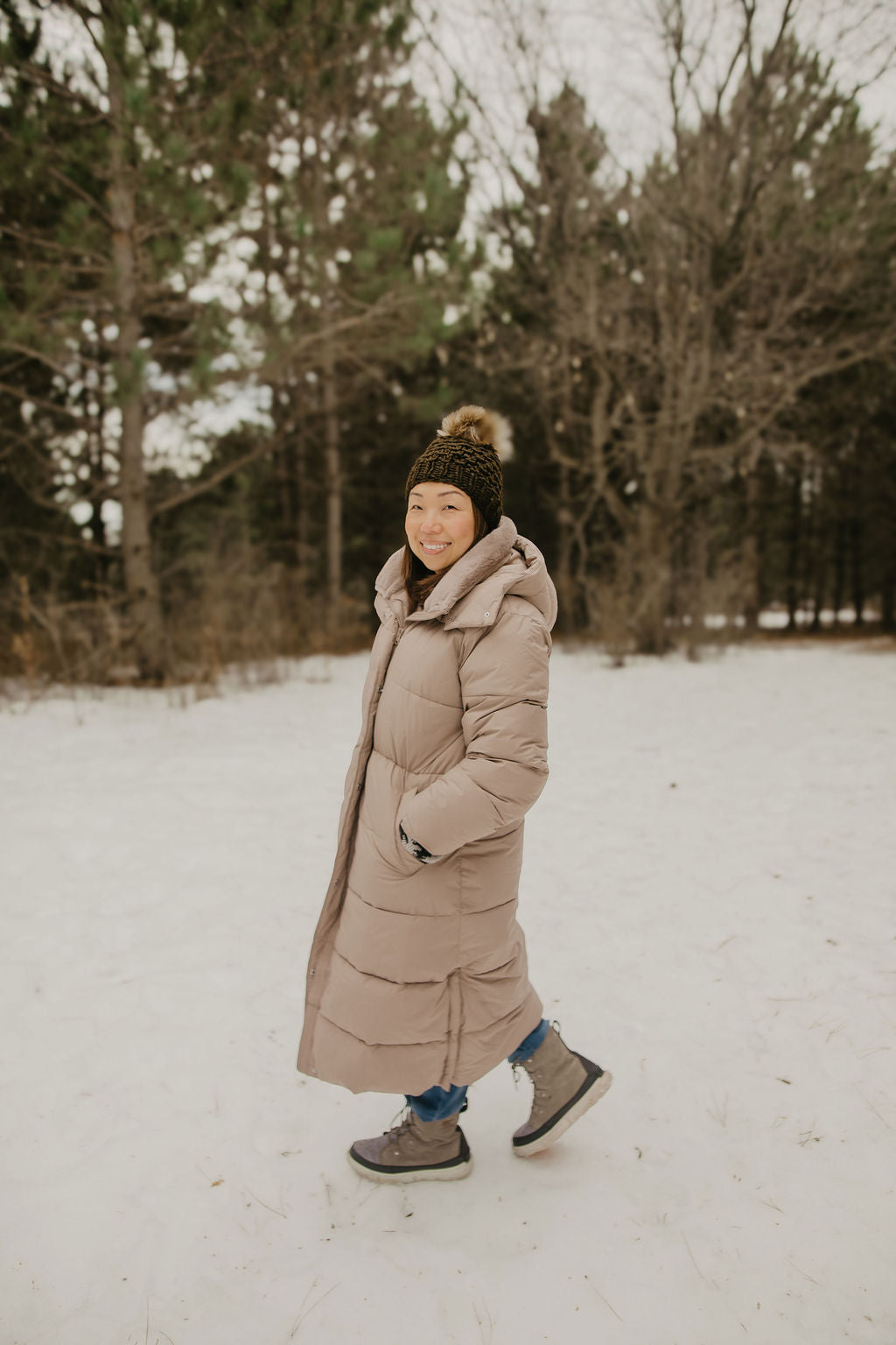 Person wearing a long beige puffer coat and green knit hat in a snowy forest