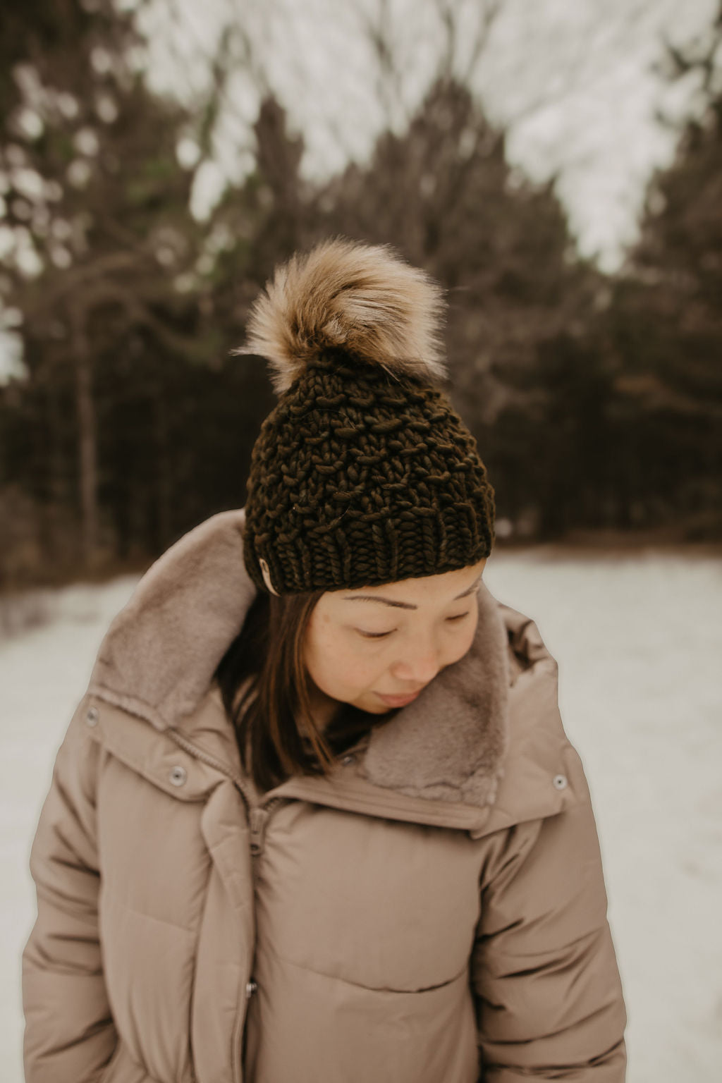 Person wearing a brown coat and green knit beanie with a fur pom-pom in a snowy forest.