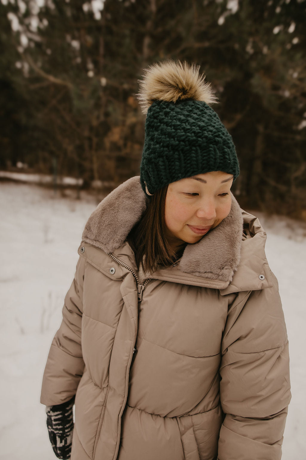 Person wearing a beige coat and green knit hat with a fur pom-pom in a snowy landscape.