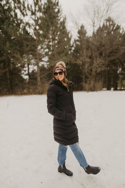 Woman wearing a long black coat, sunglasses, and a maroon knit hat in a snowy landscape with trees in the background