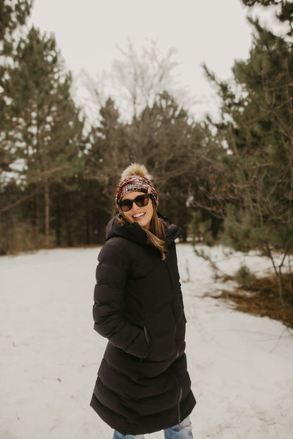 Woman in a black coat, sunglasses, and patterned beanie standing in a snowy forest.