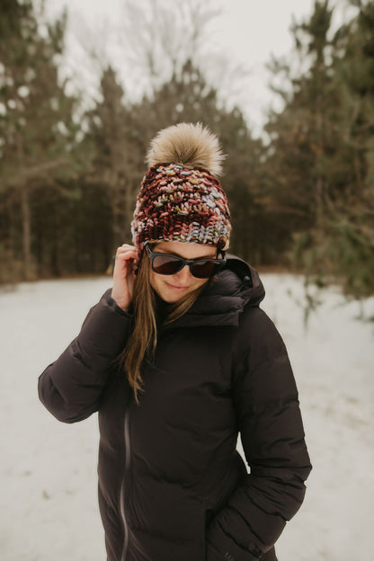 Woman wearing a colorful maroon knit hat and sunglasses and a black coat in a snowy forest