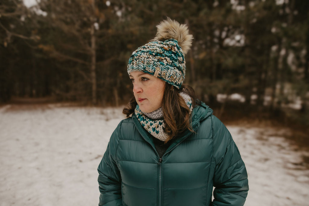 Person wearing a teal jacket and knitted hat with a pom-pom in a snowy forest setting