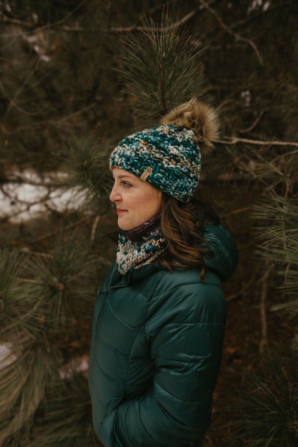Woman wearing a teal knit hat and coat standing in a forest setting