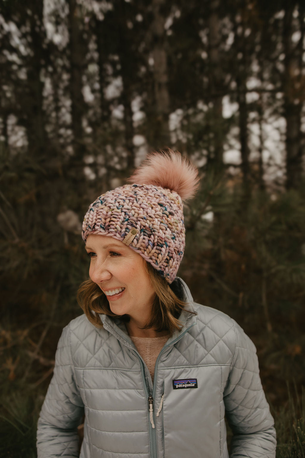 Woman wearing a pink knit hat with a pink faux fur pom pom and gray quilted jacket in a forest setting