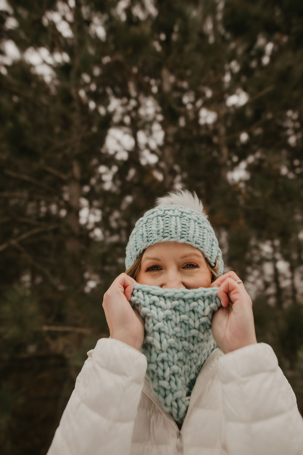 Woman wearing a green knitted hat peeks out from a knitted scarf in a snowy forest