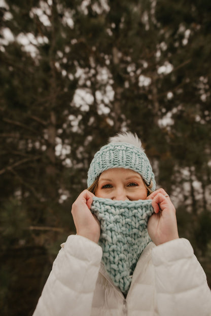 Woman wearing a green knitted hat peeks out from a knitted scarf in a snowy forest