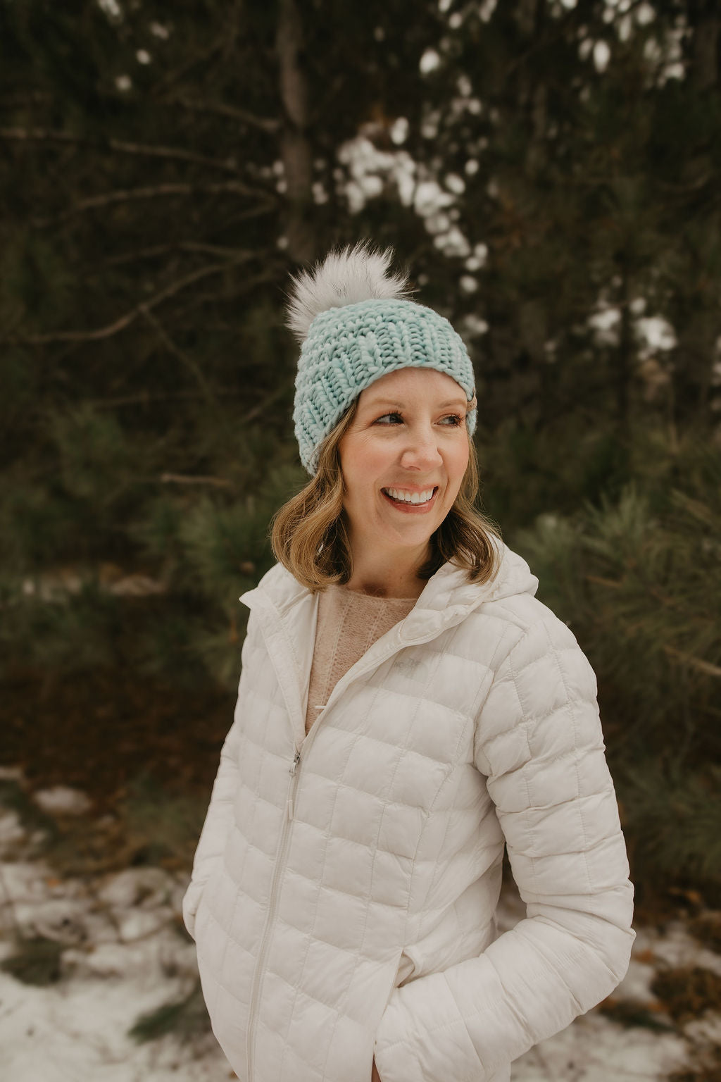 Woman wearing a white puffer jacket and light blue knit hat with a fluffy pom-pom in a snowy forest.