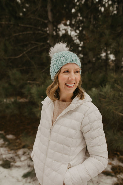 Woman wearing a white puffer jacket and light blue knit hat with a fluffy pom-pom in a snowy forest.