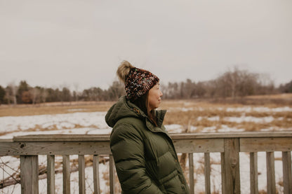 Person wearing a green coat and maroon beanie with faux fur pom pom standing on a wooden bridge over a snowy field.