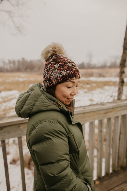 Woman wearing a green coat and maroon colorful knit beanie standing on a wooden deck with a snowy landscape in the background.