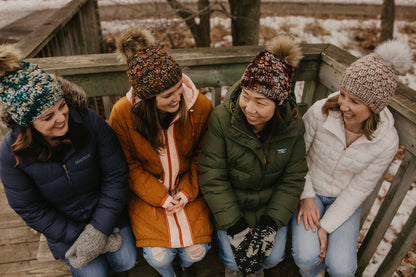 Four women sitting together on a wooden deck wearing winter clothing and knit hat with fluffy pom poms.