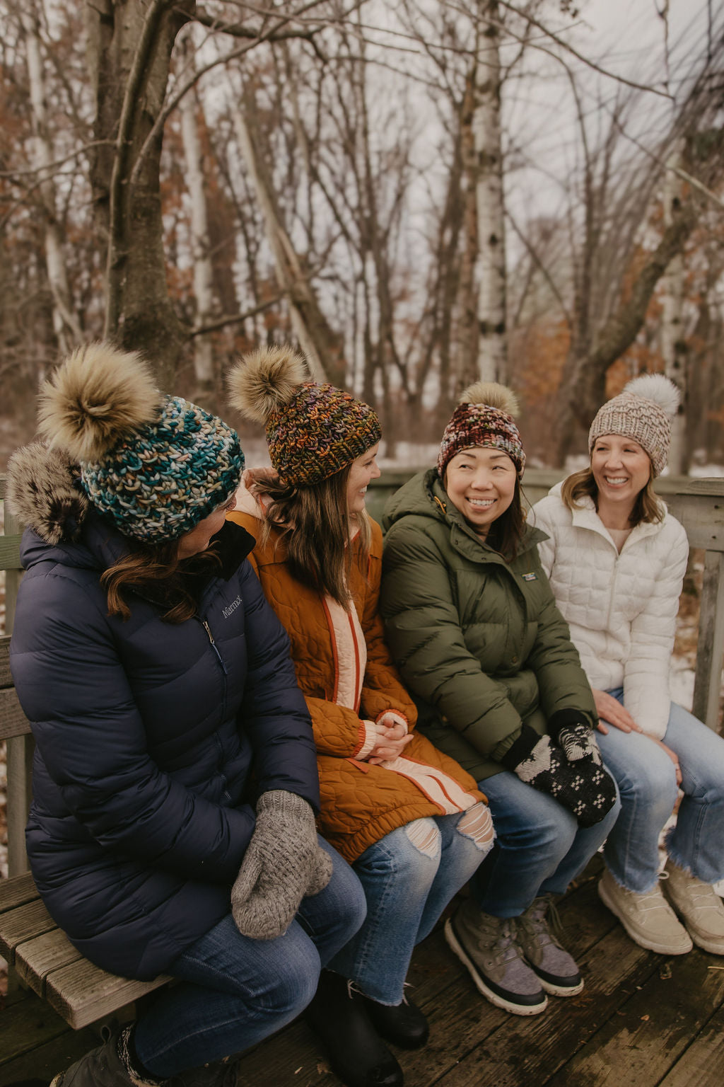 Four women sitting on a wooden bench outdoors in winter clothing and colorful knitted beanies.
