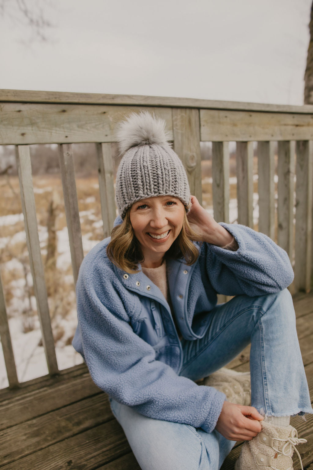 Woman wearing a gray knitted beanie and blue coat sitting on a wooden deck with a snowy landscape in the background.