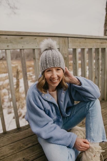 Woman wearing a gray knitted beanie and blue coat sitting on a wooden deck with a snowy landscape in the background.
