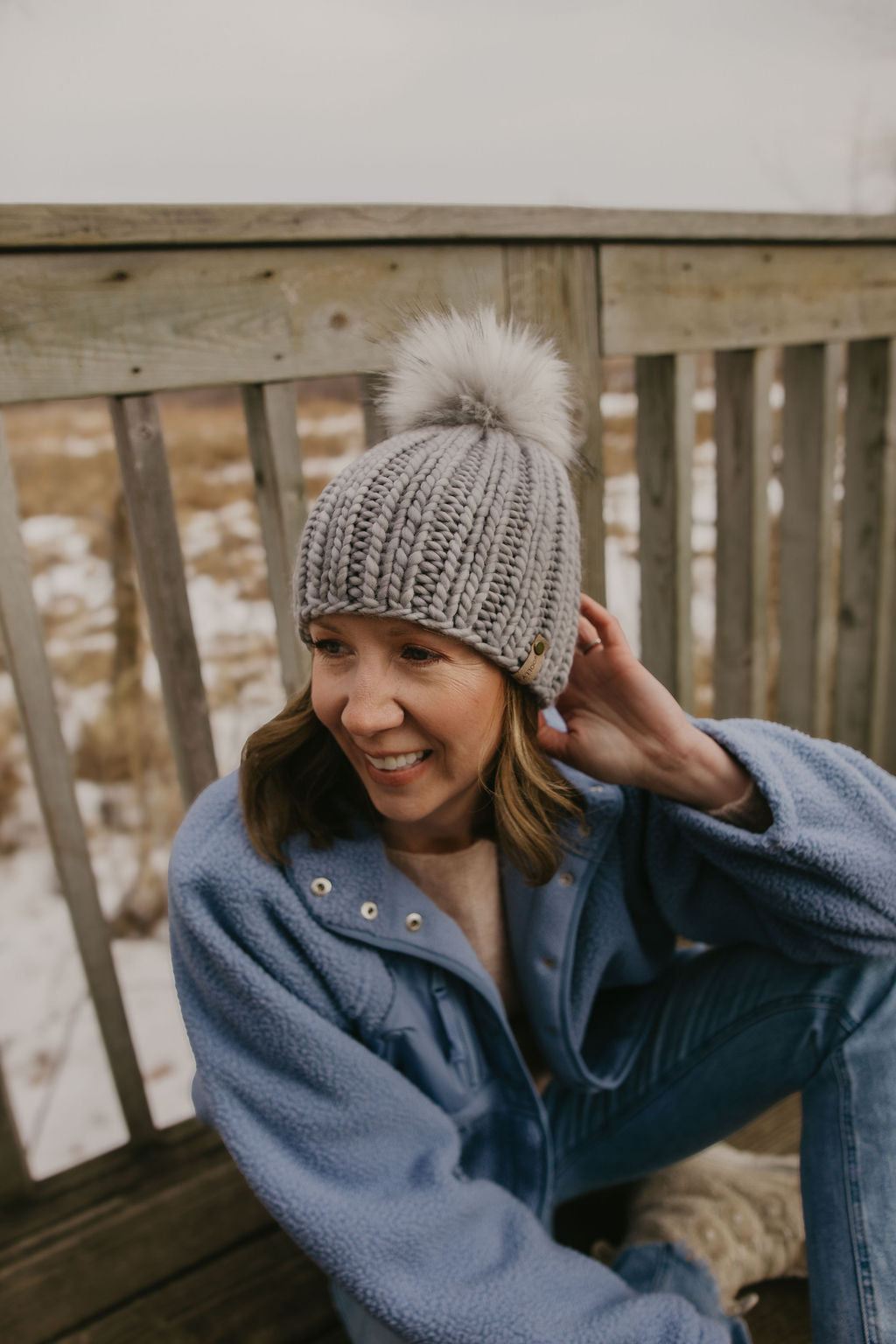 Woman wearing a gray ribbed knitted beanie with a pom-pom on a wooden deck.